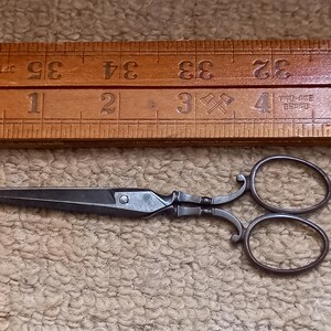 May include: Antique silver-toned scissors with a decorative design. The scissors have two rings and a small rivet on the handle. The scissors are lying on a brown surface.