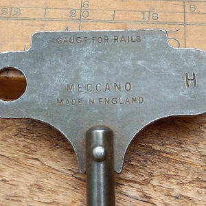 May include: A vintage metal gauge for rails, with the text "GAUGE FOR RAILS", "MECCANO", and "MADE IN ENGLAND" embossed on the surface. The gauge has a key-like shape with a handle and a hole. The metal has a worn, aged appearance.