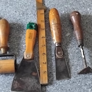 May include: A collection of five vintage hand tools with wooden handles, including a putty knife, a seam roller, a scraper, a chisel, and a triangular scraper, arranged next to a yellow wooden ruler measuring 20 centimetres in length.