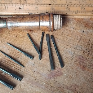 May include: A collection of vintage woodworking tools, including a wooden handled tool, a wooden measuring stick, and various metal chisels. The tools are displayed on a wooden surface, highlighting their aged appearance and craftsmanship.