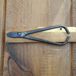 May include: Antique metal shears with curved handles and a pointed blade, resting on a wooden ruler. The shears are dark gray with signs of wear, suggesting age and use. The ruler is made of wood and has black markings.