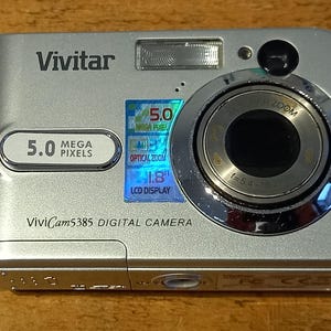 Może przedstawiać: Srebrny aparat cyfrowy Vivitar ViviCam 5385 o rozdzielczości 5,0 megapikseli i wyświetlaczu LCD o przekątnej 1,8 cala. Aparat posiada funkcję zoomu cyfrowego i srebrny obiektyw.