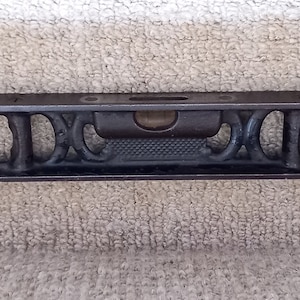 May include: A black metal level with a bubble level and two open slots on each side. The level is on a gray carpet.