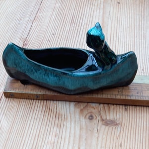 Könnte beinhalten: Ein teal-farbener Keramik-Pflanztopf in Bootsform mit einer erhabenen, stilisierten Vogelfigur an einem Ende. Der Pflanztopf hat eine glänzende Oberfläche und steht auf einer Holzoberfläche. Der bootsförmige Pflanztopf ist etwa 20 cm lang.