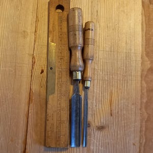 Puede incluir: Dos cinceles de mango de madera con hojas de metal, uno con una hoja negra y otro con una hoja plateada, junto a una regla de madera con marcas de latón.