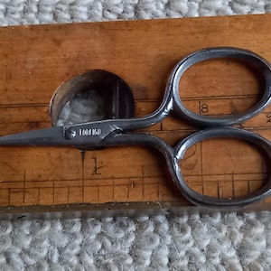 May include: A pair of small, silver-colored scissors with a wooden handle. The scissors are lying on a wooden ruler with markings in inches. The scissors have the text "1018 16" on the blade.