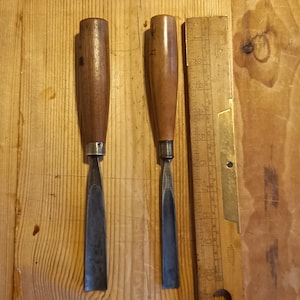 Puede incluir: Dos cinceles con mango de madera y cuchillas de metal, uno de 1,27 cm de ancho y el otro de 0,64 cm de ancho. Los cinceles están sobre una superficie de madera junto a una regla.