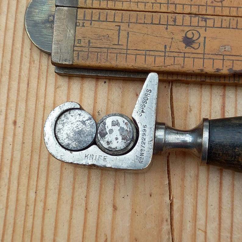 May include: A silver metal tool with a black handle for sharpening scissors and knives. The tool has the words "KNIFE" and "SCISSORS" engraved on it, along with the number "RBN 9722996".
