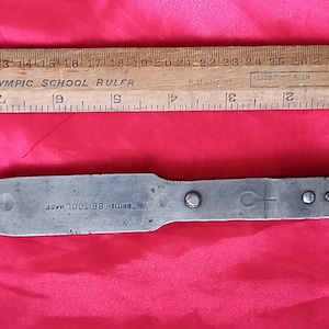 Puede incluir: Una llave metálica vintage con el texto "BRITISH BRITOOL MADE" y una regla de madera con marcas en pulgadas. La regla mide aproximadamente 30 cm de largo. La llave está sobre un fondo rojo.