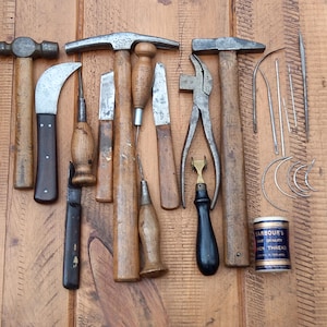 Peut inclure: Ensemble d'outils de maroquinerie anciens, comprenant des marteaux, des couteaux, des ciseaux, des pinces, des aiguilles et une petite boîte de fil. Les outils ont des manches en bois et des lames en métal, disposés sur une surface en bois.