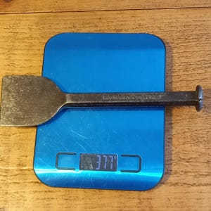 Op de afbeelding: Een vintage metalen beitel met een plat, rechthoekig blad en een cilindrische handgreep. De beitel ligt op een blauwe digitale weegschaal, die het getal 377 weergeeft. Het gereedschap lijkt gebruikt en vertoont slijtage.