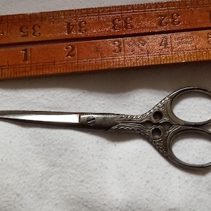 Könnte beinhalten: Eine kleine, verzierte Schere in Silber mit einem dekorativen Muster auf den Griffen. Die Schere ist etwa 10 cm lang.