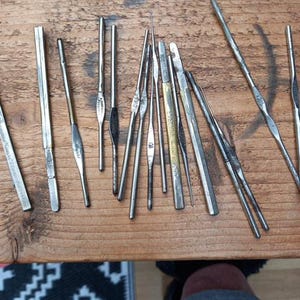 May include: A collection of vintage crochet hooks of various sizes and shapes, made of metal. The hooks are arranged on a wooden surface, showcasing their different designs and lengths. Some hooks have markings.