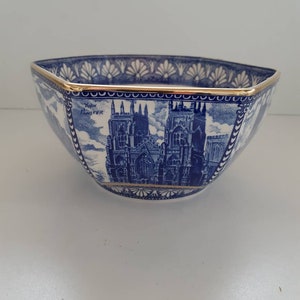 May include: A blue and white ceramic bowl with a gold rim. The bowl features a detailed illustration of a cathedral and the text "York Minster". The bowl has a hexagonal shape.
