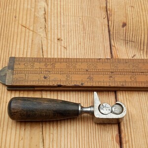 May include: A vintage metal and wood tool with a wooden handle and a metal mechanism with two circular holes. The tool is used for crimping or shaping metal.