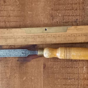 May include: A wooden-handled chisel with a metal blade. The chisel is about 6 inches long and has a brass ferrule. The chisel is lying on a wooden surface.
