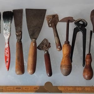 Könnte beinhalten: Eine Sammlung alter Handwerkzeuge, bestehend aus einem Hammer, vier Pinseln, zwei Spachteln, einem kleinen Metallschaber, einem speziellen Schneidewerkzeug, einer Metallzange, zwei Schraubendrehern und einem runden Maßband, angeordnet über einem Holzlineal.