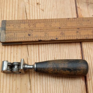 May include: A vintage metal and wood tool with a wooden handle and three metal rollers. The tool is used for shaping or smoothing materials.