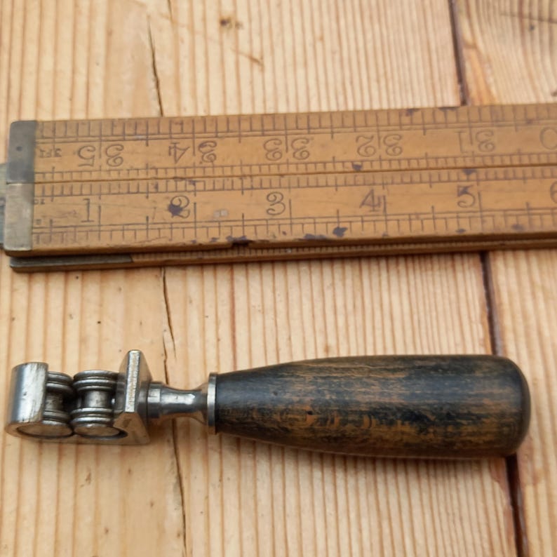 May include: A vintage metal and wood tool with a wooden handle and three metal rollers. The tool is used for shaping or smoothing materials.