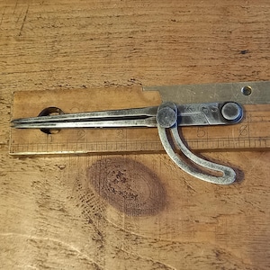 May include: Vintage metal compass on a wooden ruler. The compass is silver-toned with a curved handle and pointed tips. The ruler is made of wood and has etched markings in centimetres. The compass is positioned at the 7.6 cm mark.