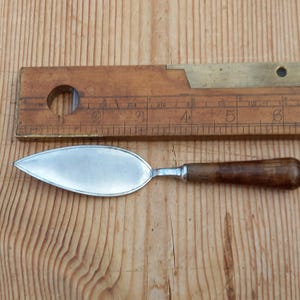 May include: A silver and wood butter knife with a pointed blade and a wooden handle. The knife is approximately 12.5 cm long.