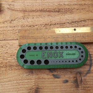 Puede incluir: Calibrador de brocas de metal verde y gris con el texto "ENOX ENGLAND" y "MADE IN". Una regla de madera está colocada encima del calibrador. El calibrador tiene múltiples agujeros de varios tamaños.