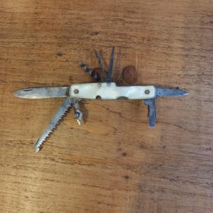 Könnte beinhalten: Ein Vintage-Multitool-Messer mit einem Griff aus Perlmutt. Das Messer verfügt über eine Hauptklinge, eine Säge, einen Korkenzieher und andere Werkzeuge. Die Metallkomponenten weisen Gebrauchsspuren auf und haben ein rustikales Aussehen. Das Messer ist geöffnet.