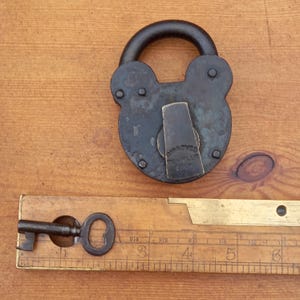 Może przedstawiać: Antyczna, ciemna metalowa kłódka z zakrzywionym uchwytem, z napisem "IMPROVED TUMBLER LOCK". Obecny jest również klucz w stylu vintage oraz drewniana linijka z oznaczeniami w calach, sugerująca przedmiot historyczny lub kolekcjonerski.