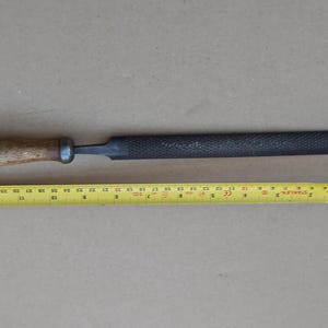 May include: A black metal file with a wooden handle. The file is approximately 25 cm long. A yellow measuring tape is visible in the background.