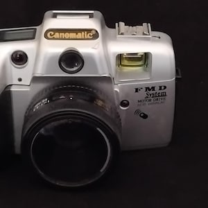May include: A silver Canomatic FMD System camera with a black lens, a built-in flash, and a black grip. The camera body features the text Canomatic, FMD System, and Motor Drive. It has a top-mounted LCD screen, a shutter button, and a viewfinder.