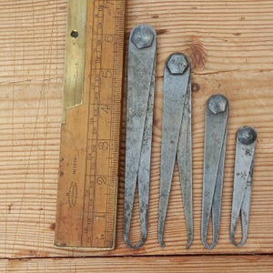 May include: Four antique metal calipers of varying sizes, with a wooden ruler in the background.