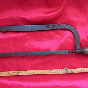 Può includere: Una sega in metallo vintage con manico in legno. La sega è lunga circa 43 cm e presenta una patina verde. È appoggiata su una superficie rossa con un righello di legno accanto.