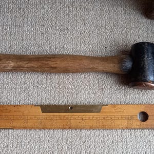 Puede incluir: Un martillo con mango de madera y cabeza de goma negra con una banda de cobre alrededor de la cabeza. También se ve una regla de madera con marcas de latón en la imagen.