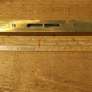 Puede incluir: Un nivel de latón con un nivel de burbuja blanco y negro y el texto "J. RABONE & SONS MAKERS BIRMINGHAM ENG" en la parte superior. El nivel está sobre una superficie de madera.