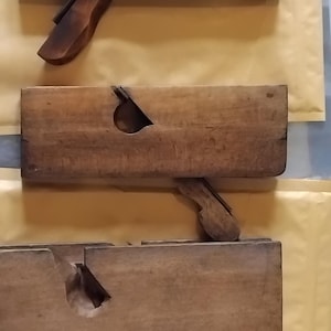 Könnte beinhalten: Drei rechteckige Handhobel aus Holz, die vertikal angeordnet sind. Jedes Werkzeug besteht aus einem glatten, verwitterten braunen Holzkörper mit einem zentralen, schrägen Schlitz, der eine Metallklinge und einen Holzkeil enthält. Die Maserung ist sichtbar.