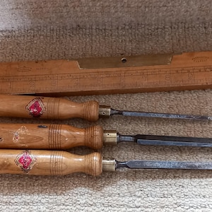 Może przedstawiać: Trzy dłuta z drewnianymi uchwytami i metalowymi ostrzami. Uchwyty są oznaczone czerwonym diamentowym logo. W tle znajduje się drewniana linijka z mosiężnymi oznaczeniami.