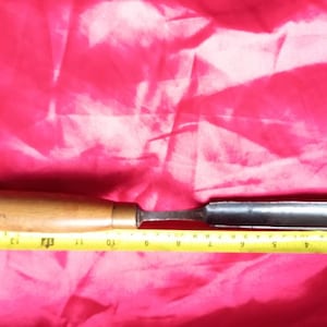 Puede incluir: Un cincel de mango de madera con una hoja de metal, que mide 28 cm de largo, sobre un fondo de tela rosa.