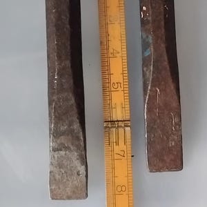 Op de afbeelding: Twee verweerde, donkergrijze metalen beitels met platte, afgeschuinde randen, verticaal geplaatst. Een gele houten vouwmeter ligt tussen de twee gereedschappen en toont maten in centimeters, waarbij de gereedschappen ongeveer 23 cm lang zijn.