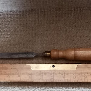 May include: A wooden handled chisel with a metal blade, measuring 10 inches long, resting on a wooden ruler with brass markings.