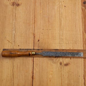 May include: A wooden-handled chisel with a metal blade. The chisel is resting on a wooden surface.