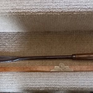 May include: A long, thin, metal tool with a wooden handle. The tool is about 12 inches long and has a flat, rectangular blade. The handle is brown and has a slight curve. The tool is lying on a white, textured surface.