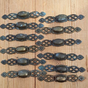 May include: Ten antique brass drawer pulls with an ornate design. Each pull has a rounded oval shape in the center and a decorative, intricate pattern around the edges.