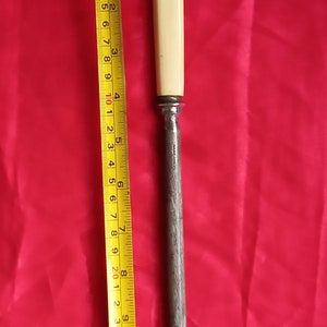 May include: A vintage steel knife sharpener with a cream-coloured handle. The sharpener is approximately 25 cm long, next to a yellow measuring tape. The background is a solid red colour.