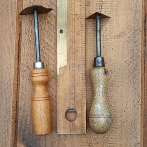 May include: Two antique metal and wood tools with wooden handles and metal blades. A vintage ruler is positioned between the tools. The tools are set against a wooden background.