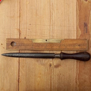 May include: A black metal awl with a wooden handle. The awl is about 10 inches long and has a sharp point. The handle is about 4 inches long and is made of dark wood.
