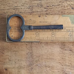 Könnte beinhalten: Ein dunkler Metallschlüssel mit rundem Griff und rechteckigem Schaft, der auf einem Holzlineal liegt. Das Lineal zeigt Zentimetermessungen und den Text "RABONE & SONS BIRMINGHAM MADE IN ENGLAND". Der Schlüssel ist abgenutzt.