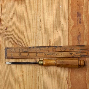 Peut inclure: Un ciseau à bois vintage avec une virole en laiton et un manche en bois, à côté d'une règle pliante. Le ciseau a une lame en métal foncé. La règle est en bois et porte des marques noires.
