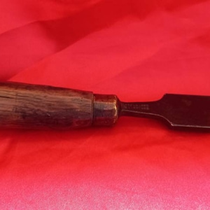 Peut inclure: Un ciseau à bois avec une lame en métal. Le manche est brun foncé et la lame est noire. Le ciseau mesure environ 25 cm de long.