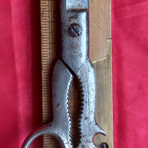 May include: Antique metal scissors with a serrated edge and bottle opener. The scissors are marked "ITALY" and are next to a wooden ruler. The scissors are on a red background. The ruler has measurements.