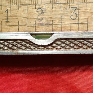 Puede incluir: Un nivel de metal plateado con un nivel de burbuja verde. El nivel mide aproximadamente 5 cm de largo y tiene un patrón de diamante en la parte superior.